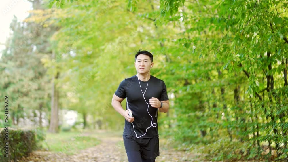 Young adult asian runner with headphones and smartphone jogging in urban city park. Handsome man in a sports suit likes to run in the fresh air while listening to his favorite music. Healthy lifestyle Young adult asian runner with headphones and smartphone jogging in urban city park. Handsome man in a sports suit likes to run in the fresh air while listening to his favorite music. Healthy lifestyle