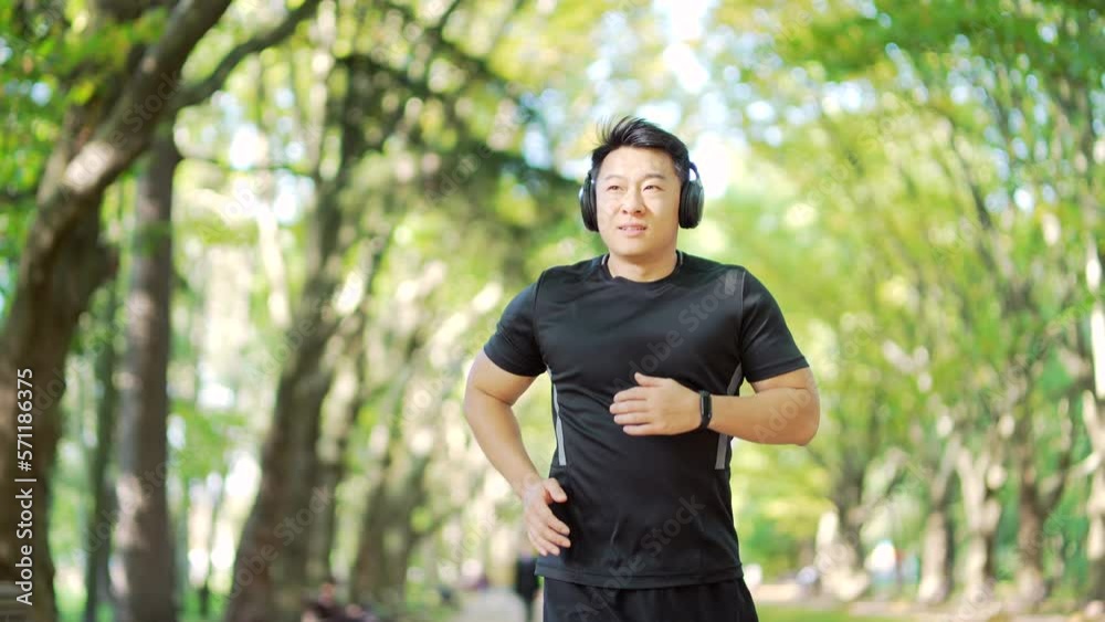 Young adult asian runner with headphones jogging in an urban city park. Handsome male in a tracksuit enjoys jogging outdoors while listening to his favorite music. Concept sport and healthy lifestyle Young adult asian runner with headphones jogging in an urban city park. Handsome male in a tracksuit enjoys jogging outdoors while listening to his favorite music. Concept sport and healthy lifestyle