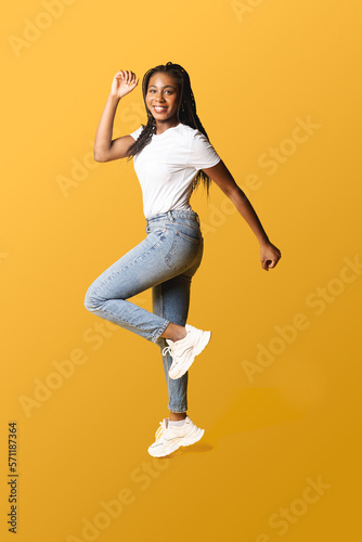 Full length photo of happy young 20s african woman with pigtails in white basic t-shirt and jeans jumping high, inspired by sale or best promotion, copy space, isolated on yellow