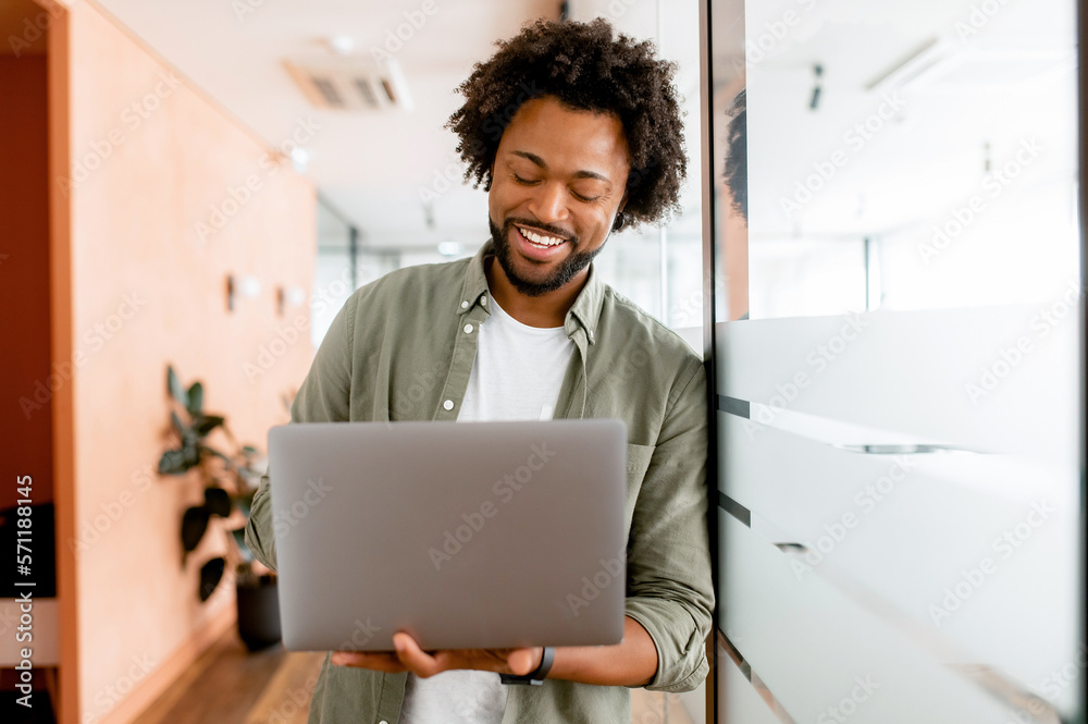 Curly young african-american businessman standing in office hallway and using laptop for office work, male employee checks mail, conducts business correspondence with customers - obrazy, fototapety, plakaty 