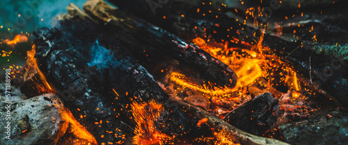 Vivid smoldered firewoods burned in fire close-up. Atmospheric background with orange flame of campfire and blue smoke. Warm full frame image of bonfire. Glowing embers in air. Bright sparks in bokeh