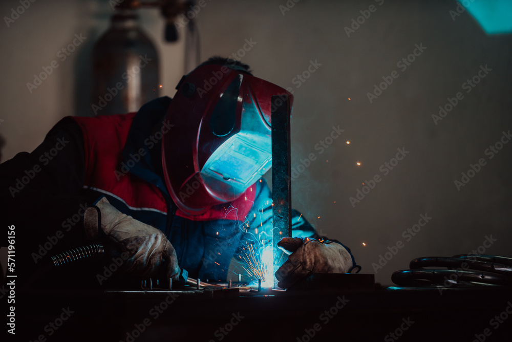 Profesional welder in protective uniform and mask welding metal pipe on the industrial table with other workers behind in the industrial workshop. Profesional welder in protective uniform and mask welding metal pipe on the industrial table with other workers behind in the industrial workshop.