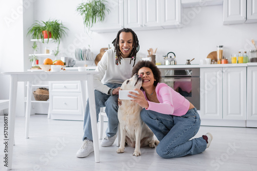 Smiling african american couple taking selfie with labrador in kitchen.
