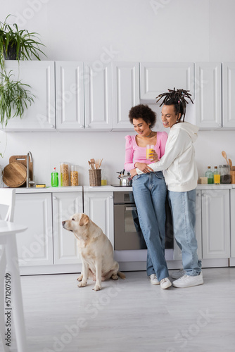 Smiling african american couple with orange juice looking at labrador in kitchen.