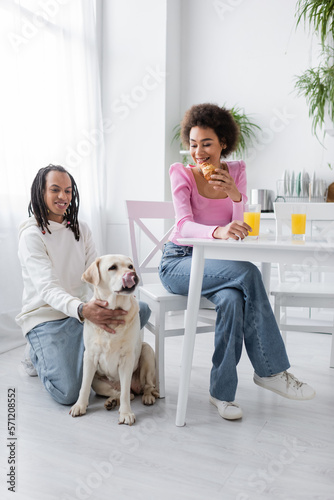 Smiling african american woman holding croissant near boyfriend and labrador in kitchen.