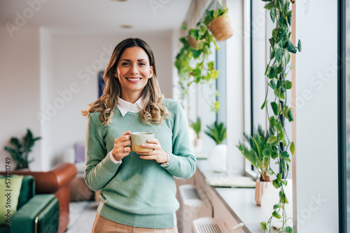 Portrait of a pretty businesswoman, holding a cup of coffee, standing at the home office, smiling. © bnenin