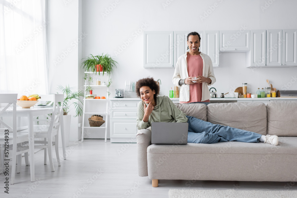 Positive african american couple with laptop and coffee looking at camera in kitchen.