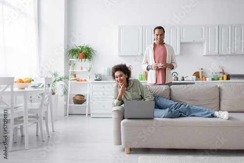 Positive african american couple with laptop and coffee looking at camera in kitchen.