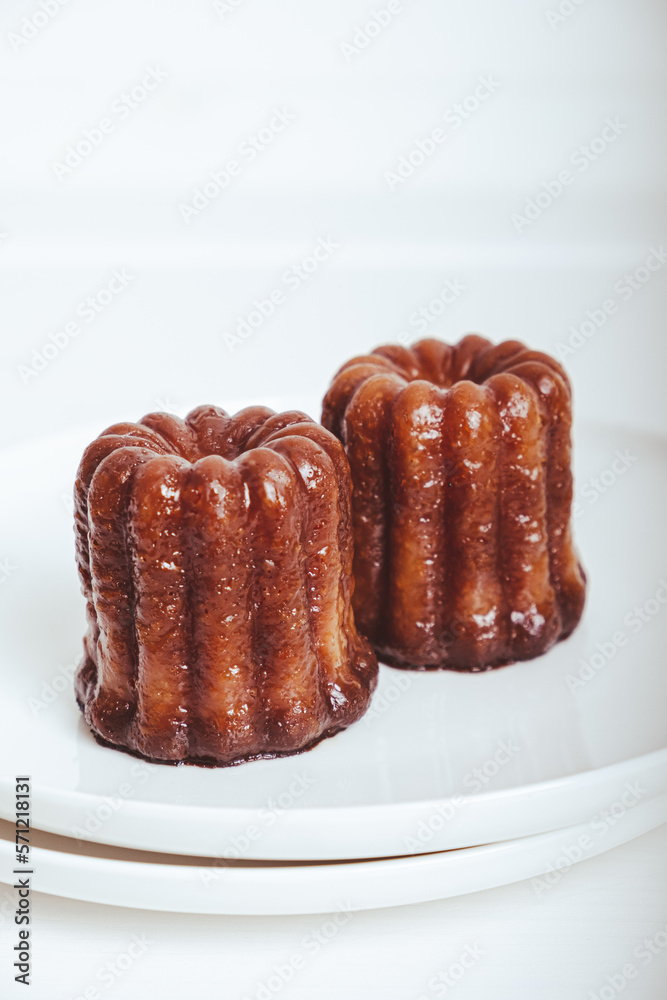 Delicious freshly baked canele, rum and vanilla flavored french pastry with caramelized crust on white plate against white background, close up. A specialty of the Bordeaux region of France. Delicious freshly baked canele, rum and vanilla flavored french pastry with caramelized crust on white plate against white background, close up. A specialty of the Bordeaux region of France.