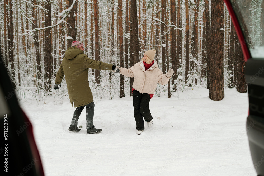 Happy senior couple in winterwear having fun and dancing in pinetree forest while enjoying weekend in front of their car with open door
