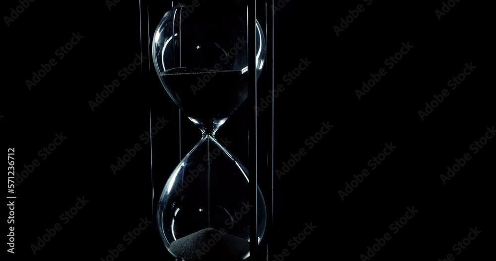 Rotation of sand timer clock with sand flowing from through hourglass on black background., Tilt down
