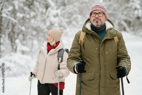 Mature man in winterwear strolling with trekking sticks in the forest covered with snow while his wife standing on background
