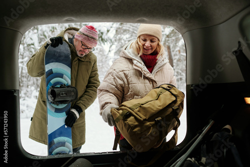 Senior woman putting backpack in car trunk while her husband with snowboard standing next to her during their weekend trip