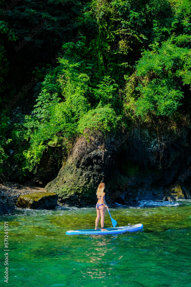Fototapeta premium SUP Stand up paddle board. Blond girl in a swimsuit sailing on paddle board in the green sea near stone beach. Woman on summer holidays vacation lifestyle. Mtsvane Kontskhi Beach, Batumi, Georgia.