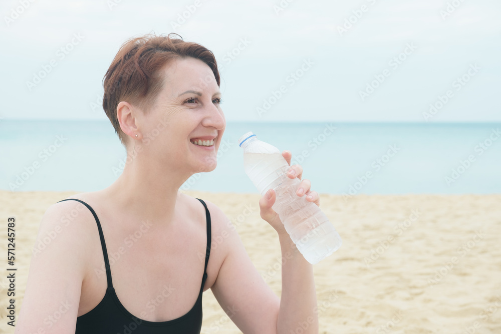 middle-aged woman with a short haircut drinks water from a plastic bottle on the beach. A woman in a bathing suit drinks water. The need to drink fluids while in the heat. Rest on the sea coast. middle-aged woman with a short haircut drinks water from a plastic bottle on the beach. A woman in a bathing suit drinks water. The need to drink fluids while in the heat. Rest on the sea coast.
