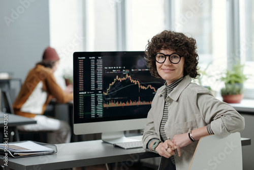 Young smiling businesswoman or crypto analyst in casualwear sitting by workplace with computer monitor and graphic data on screen