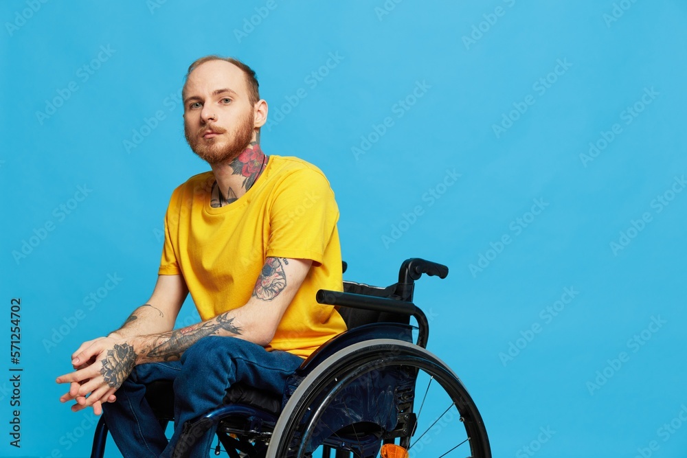 A man in a wheelchair problems with the musculoskeletal system looks at the camera in a t-shirt with tattoos on his arms sits on a blue studio background, full life, real person, health concept