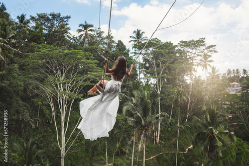 Woman wearing white dress swinging on rope swings with beautiful view on rice terraces and palm trees in the Bali Island
