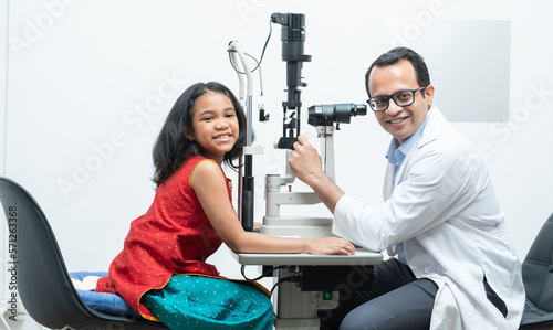 Indian middle aged professional ophthalmologist or optometrist man work at optometry clinic, using optometry instruments little girl patient to check eyesight and eyes vision. smile at camera