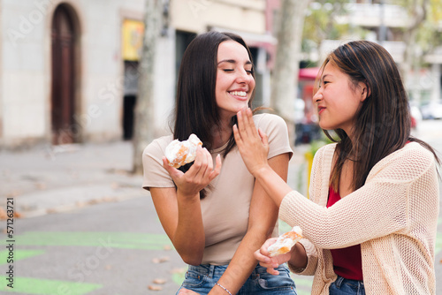 two young friends laughing and having fun while sharing some sweet buns sitting in a city park, concept of friendship and love between people of the same sex