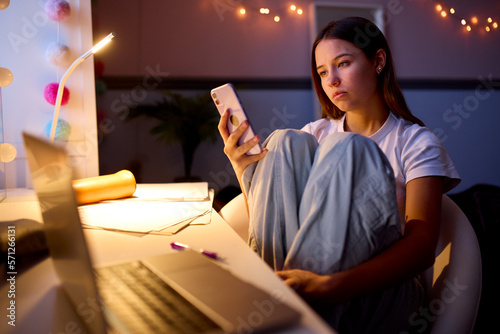 Worried Teenage Girl Sitting At Desk In Bedroom At Home Looking At Mobile Phone At Night photo