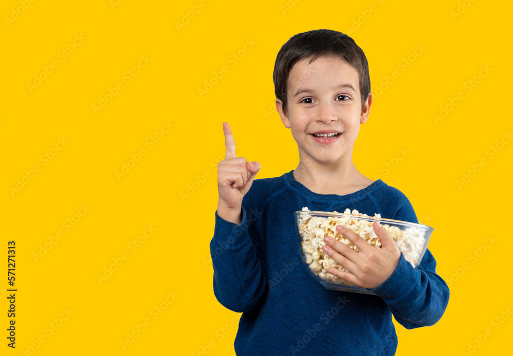 Dark haired little child eating popcorn snack isolated yellow background very happy pointing with hand and finger to the side Dark haired little child eating popcorn snack isolated yellow background very happy pointing with hand and finger to the side
