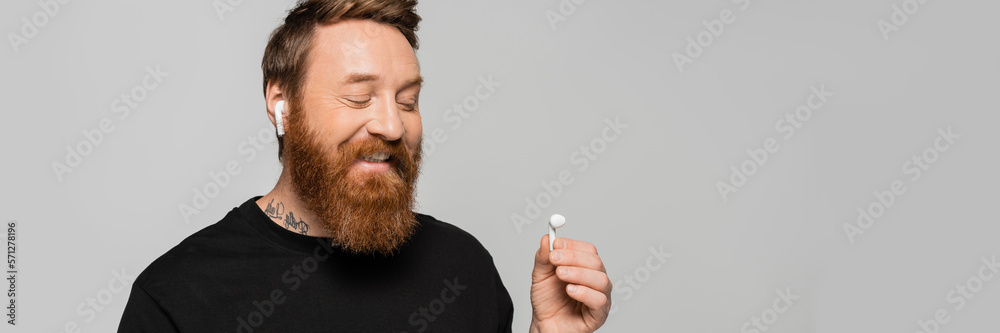 Obraz premium joyful bearded man in black t-shirt listening music and looking at wireless earphone isolated on grey, banner.