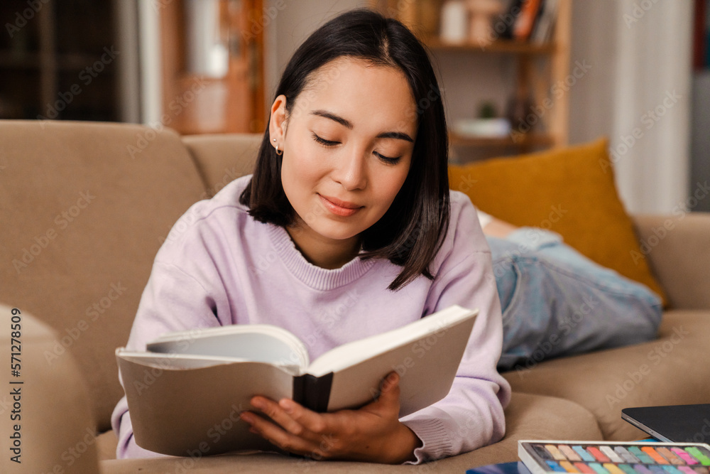 Smiling asian woman reading book while laying on couch Smiling asian woman reading book while laying on couch