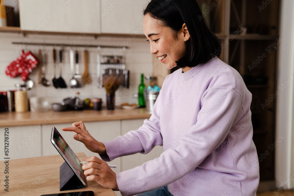 Cheerful asian woman working on tablet while sitting in kitchen Cheerful asian woman working on tablet while sitting in kitchen