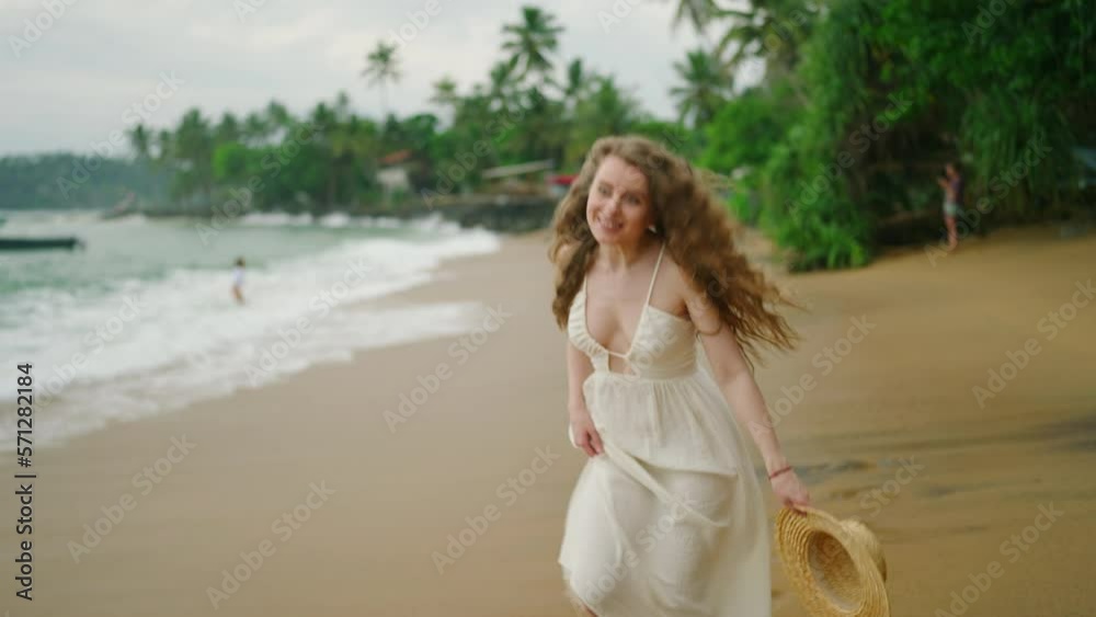 Young curly caucasian woman in white dress with straw hat in hands walks along the beach touches long hair enjoys view and smiles. Happy pretty female walking by the sea. Carefree girl enjoys summer Young curly caucasian woman in white dress with straw hat in hands walks along the beach touches long hair enjoys view and smiles. Happy pretty female walking by the sea. Carefree girl enjoys summer