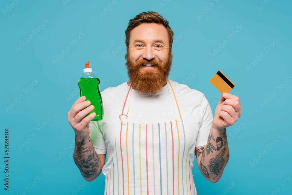 happy bearded man with dishwashing gel and credit card smiling at camera isolated on blue.