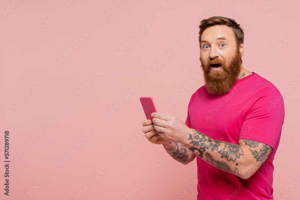 excited bearded man with open mouth using cellphone and looking at camera isolated on pink.