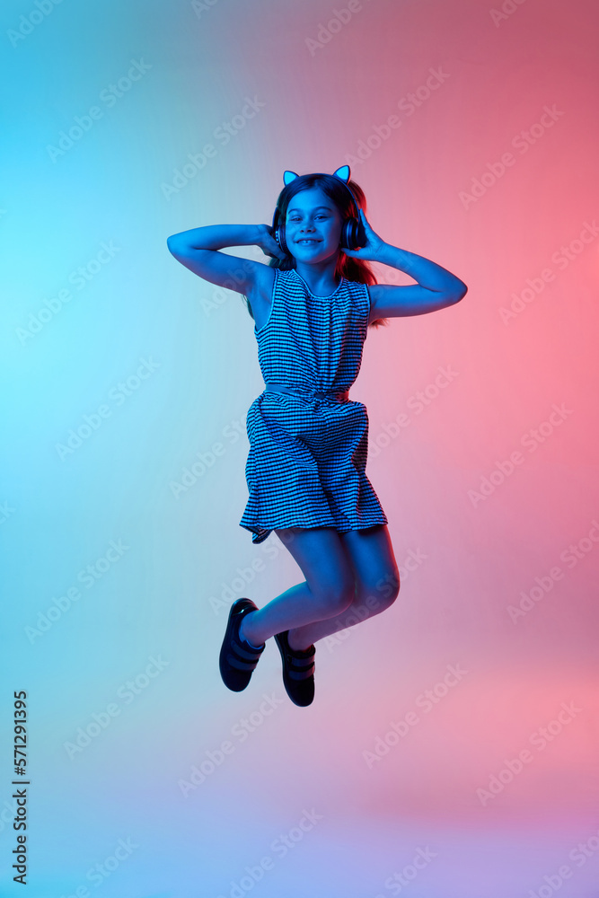 Little beautiful girl, child listening to music in headphones and jumping over gradient blue pink studio background in neon light. Childhood, emotions, fun, fashion, lifestyle, facial expression