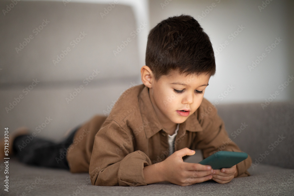 Busy serious european small kid plays on phone, watches video, lies on sofa in living room interior Busy serious european small kid plays on phone, watches video, lies on sofa in living room interior