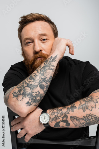 pensive tattooed man in black t-shirt and wristwatch sitting on chair and looking at camera isolated on grey.