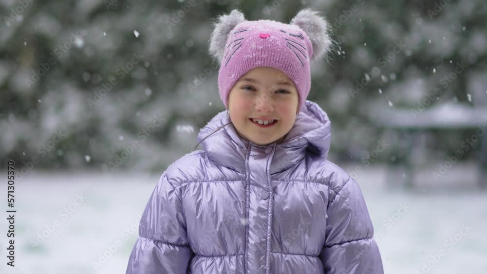 Medium close-up portrait of cheerful girl in warm clothes standing against pine wood on snowy winter day smiling at camera slow motion. Snowstorm and blizzard in winter season Medium close-up portrait of cheerful girl in warm clothes standing against pine wood on snowy winter day smiling at camera slow motion. Snowstorm and blizzard in winter season