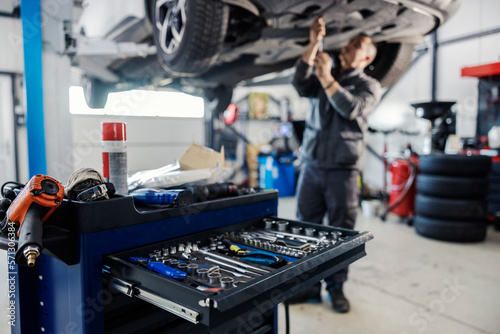 Selective focus on mechanic's toolbox with tools in drawer with auto-mechanic fixing car in blurry background at workshop.