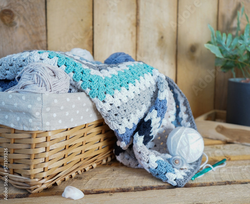 white, grey, blue granny square blanket with woolen balls in an white textile basket and crochet hooks on wooden ground