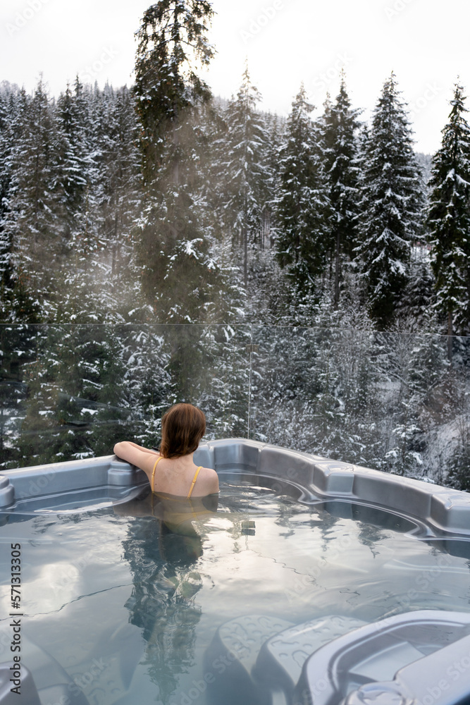 Vertical image of a young woman sitting on back in a hot tub bath and enjoying the view of a snowy forest. The concept of relaxation and water treatments. Idea of escape and recreation on nature Vertical image of a young woman sitting on back in a hot tub bath and enjoying the view of a snowy forest. The concept of relaxation and water treatments. Idea of escape and recreation on nature