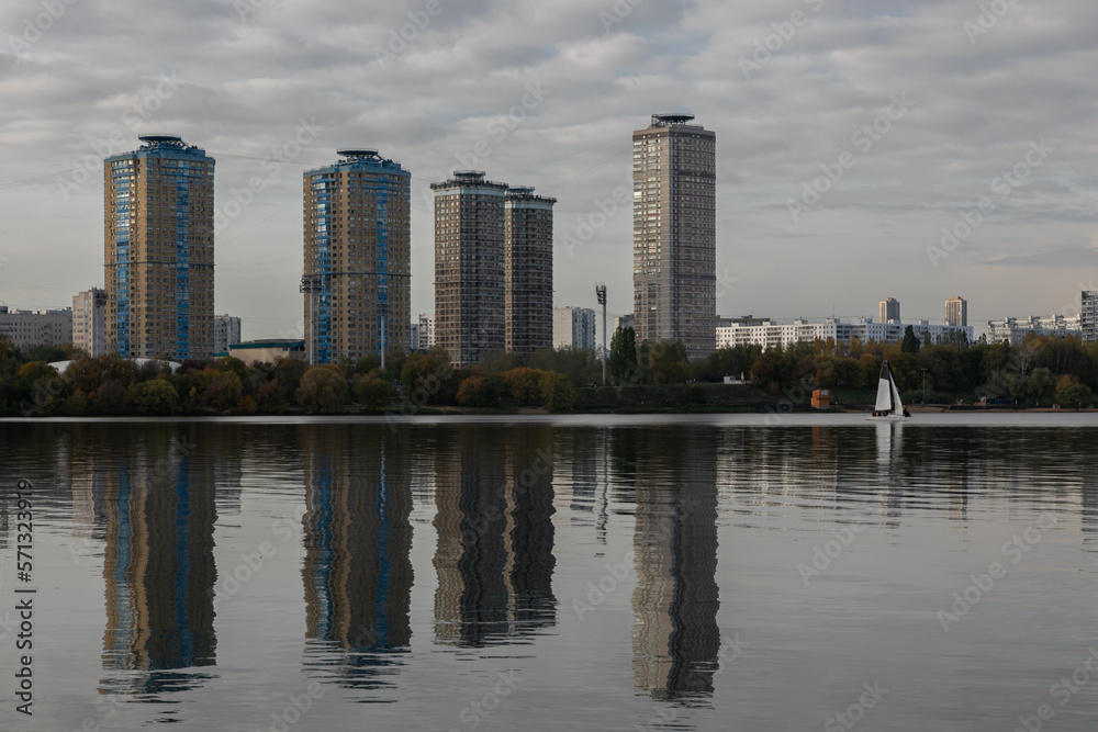 Landscape of the city lake. Strogino beach, Moscow. A city park along the embankment, several tall apartment buildings are reflected in the water surface. A lone sailboat goes down the river