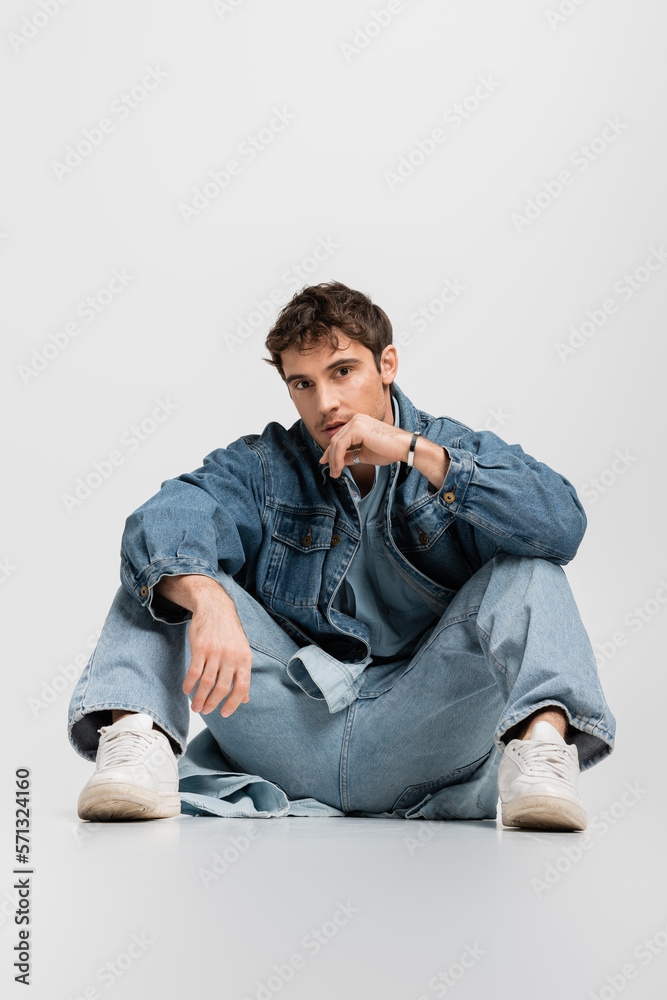 full length of stylish man in denim jacket and jeans sitting and looking at camera on grey. full length of stylish man in denim jacket and jeans sitting and looking at camera on grey.