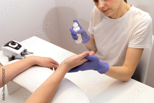 Nails master spraying, sanitizing client's hands by antiseptic from white bottle at work station. Beautician wears gloves, holding woman's hand over table. Hardware nail machine is beside. Horizontal