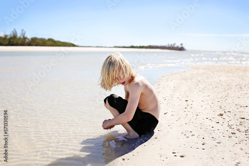 Little Boy with Long Hair Picking Up Seashells on Beach at Ocean photo
