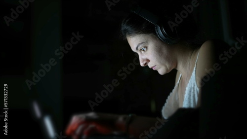 Focused woman typing on laptop keyboard in the dark wearing headphones. Person working in the dark. Modern lifestyle