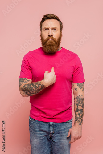 displeased bearded man in trendy t-shirt pointing with finger at himself isolated on pink.