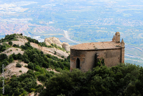 An old chapel in yellow tones, seen from behind, overlooking the valley, lost in the midst of a mountain of stones and green vegetation.