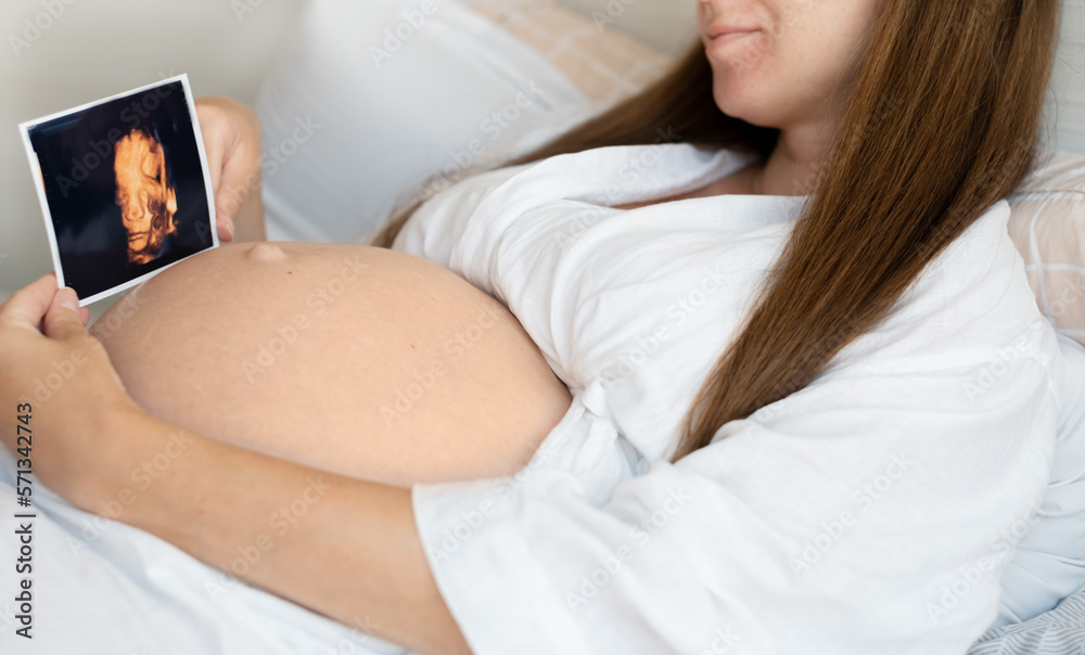 Unrecognizable pregnant woman wearing robe at home caressing,stroking belly, baby bump, holding ultrasound sonogram photo.Cropped, close-up shot.Happy,natural,calm, healthy pregnancy. Unrecognizable pregnant woman wearing robe at home caressing,stroking belly, baby bump, holding ultrasound sonogram photo.Cropped, close-up shot.Happy,natural,calm, healthy pregnancy.