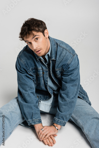 stylish man in trendy denim outfit sitting and looking at camera on grey.