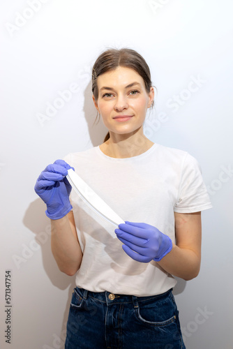Caucasian manicurist, nail technician holds file in hands, wearing gloves. White smiling manicure master stands near gray wall in spa salon. Soft, safe beauty nail care, treatment. Vertical plane photo