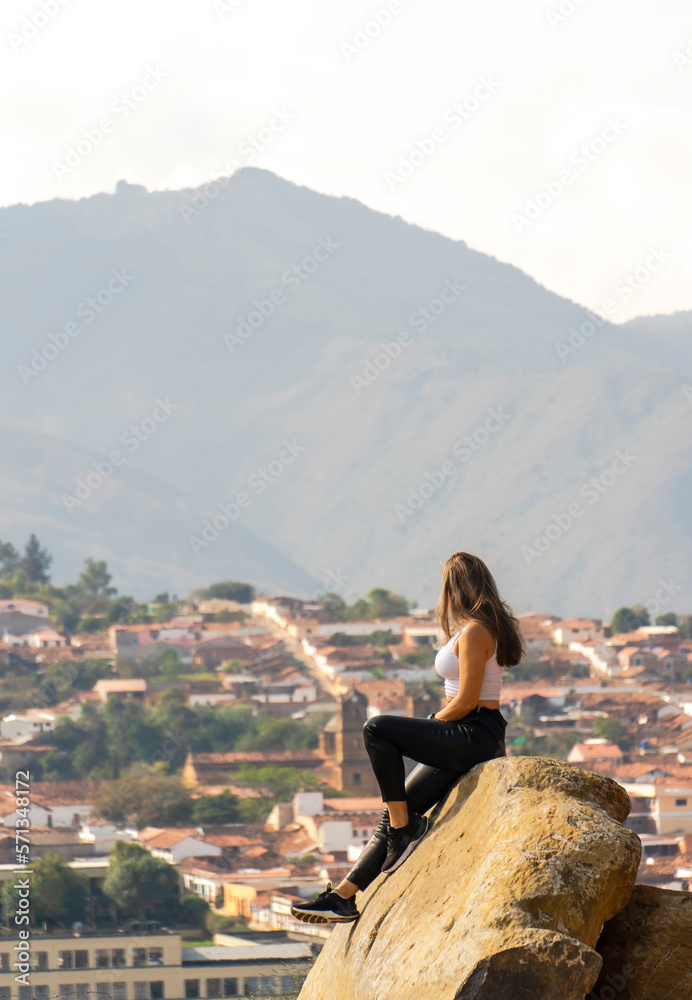 beautiful woman sitting and standing on a huge rock with the city or town in the background dressed in black pants and white blouse